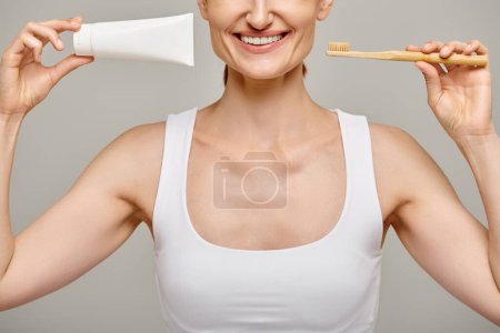 cropped view of joyful woman holding toothpaste and toothbrush and smiling at camera on grey