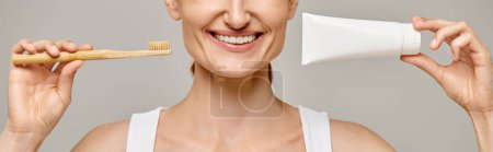 cropped banner of joyful woman holding toothpaste and toothbrush and smiling at camera on grey