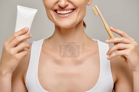 cropped view of cheerful woman holding toothpaste and toothbrush and smiling at camera on grey