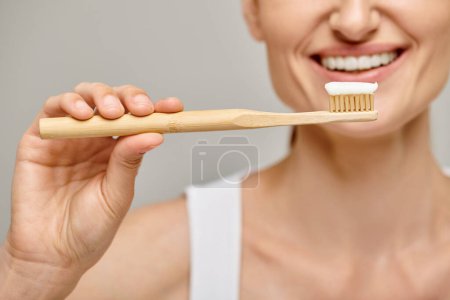 cropped view of cheerful woman holding toothbrush with toothpaste and smiling at camera on grey