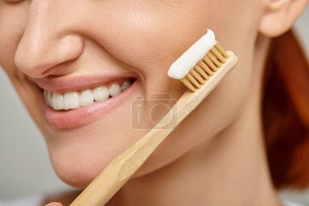 cropped view of positive woman holding toothbrush with toothpaste and smiling at camera on grey