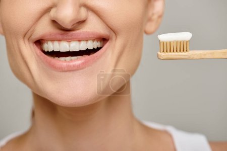 cropped view of jolly woman holding toothbrush with toothpaste and smiling at camera on grey