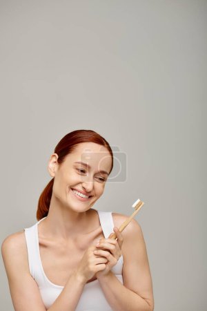 Frau mit roten Haaren schaut auf Bambuszahnbürste mit Zahnpasta vor grauem Hintergrund, Zahnpflege