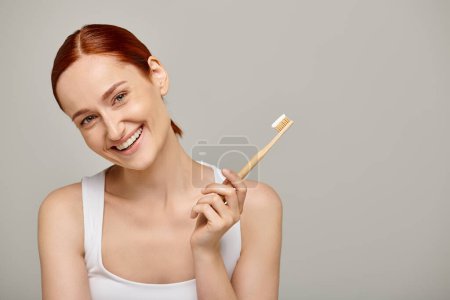 femme heureuse aux cheveux roux tenant la brosse à dents en bambou avec dentifrice sur fond gris, dentaire