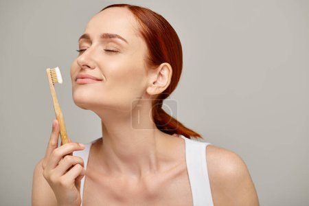 femme aux cheveux roux tenant une brosse à dents en bambou avec dentifrice sur fond gris, soins dentaires