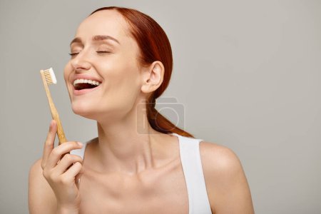 femme excitée aux cheveux roux tenant la brosse à dents en bambou avec dentifrice sur fond gris, soins dentaires