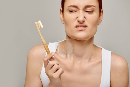 femme mécontente aux cheveux roux regardant la brosse à dents en bambou avec du dentifrice sur fond gris, dentaire