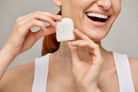 cropped view of cheerful redhead woman holding dental floss case and smiling on grey background