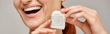 cropped banner of positive redhead woman holding dental floss case and smiling on grey background