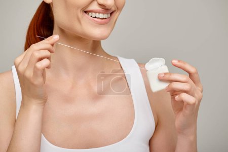 cropped view of happy redhead woman holding dental floss in case and smiling on grey background