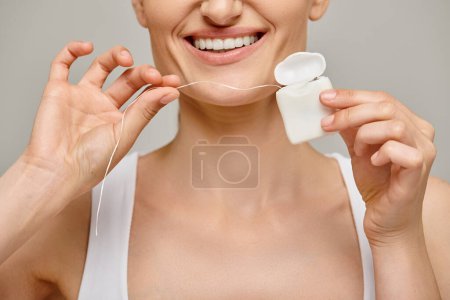 Téléchargez les photos : Vue recadrée d'une femme rousse heureuse tenant un fil dentaire et un boîtier blanc, souriant sur fond gris - en image libre de droit