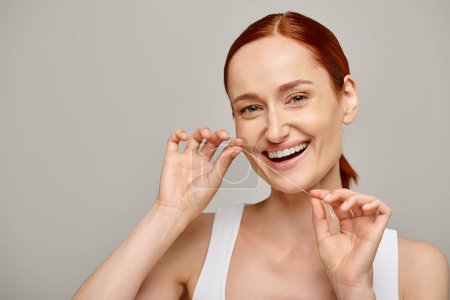 happy redhead woman holding dental floss and smiling on grey background,  promoting oral hygiene