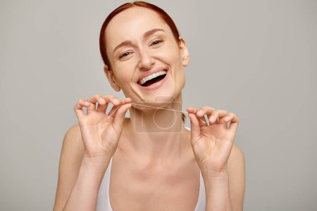 aufgeregte rothaarige Frau, die Zahnseide in der Hand hält und auf grauem Hintergrund lächelt und Mundhygiene fördert