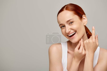 modelo pelirroja excitada sosteniendo hilo dental y sonriendo sobre fondo gris, promoviendo la higiene bucal