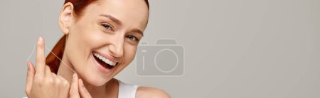 modelo pelirroja excitada sosteniendo hilo dental y sonriendo sobre fondo gris, banner de higiene oral