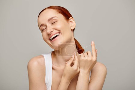 mujer alegre y pelirroja sosteniendo hilo dental y sonriendo sobre fondo gris, higiene bucal