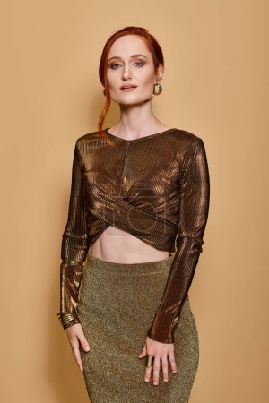 Elegant redhead woman in her 30s posing in golden attire and accessories on beige backdrop