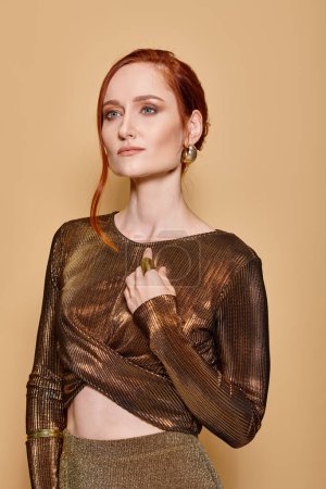 Elegant and redhead woman in her 30s posing in golden attire and accessories on beige backdrop