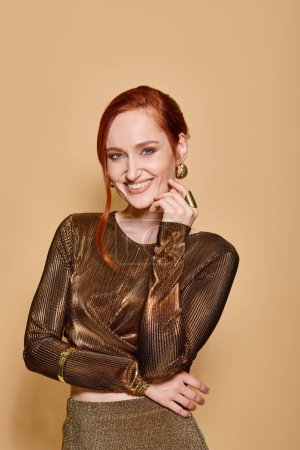 happy and redhead woman in her 30s posing in golden attire and accessories on beige backdrop