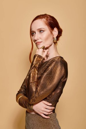 nachdenkliche rothaarige Frau in ihren Dreißigern posiert in eleganter Kleidung mit goldenen Accessoires auf beigem Hintergrund