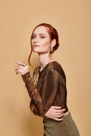 graceful redhead woman in her 30s posing in elegant attire with golden accessories on beige backdrop
