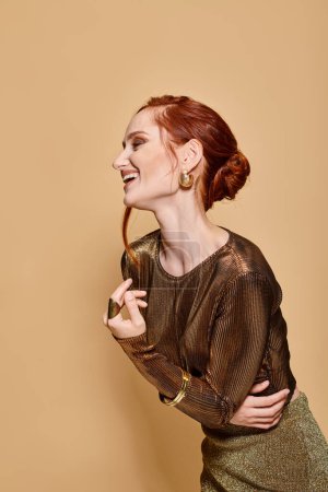 redhead woman in her 30s posing in golden attire and accessories and smiling on beige backdrop