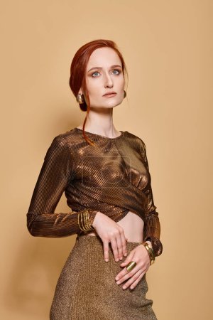 graceful redhead woman in her 30s posing in trendy attire with golden accessories on beige backdrop