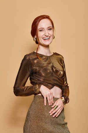 cheerful and redhead woman in her 30s posing in golden attire and accessories on beige backdrop