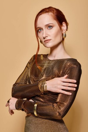 redhead woman in her 30s posing in glamorous attire with golden accessories on beige backdrop