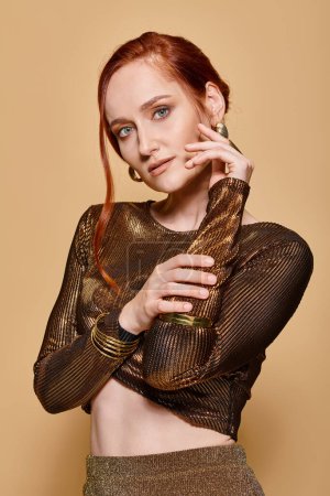 redhead woman in her 30s posing in stylish outfit and golden accessories with hand near face