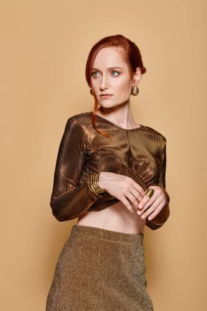 redhead woman in her 30s posing in stylish attire and golden accessories while looking away on beige