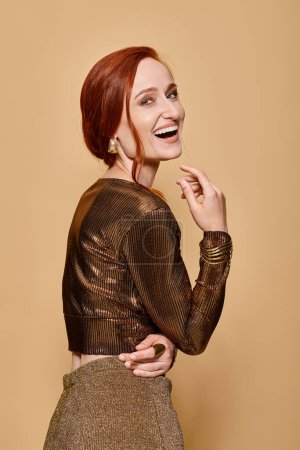 Téléchargez les photos : Joyeuse rousse femme dans la trentaine posant en tenue tendance et accessoires dorés sur fond beige - en image libre de droit