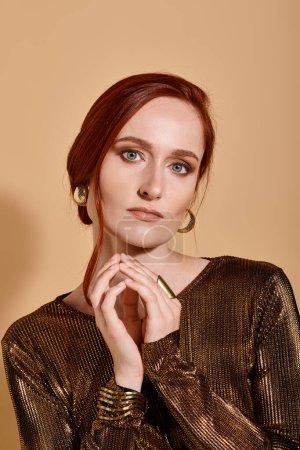 charming redhead woman in 30s posing in stylish outfit and golden accessories on beige backdrop