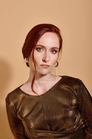 attractive redhead woman in 30s posing in stylish outfit and golden accessories on beige backdrop