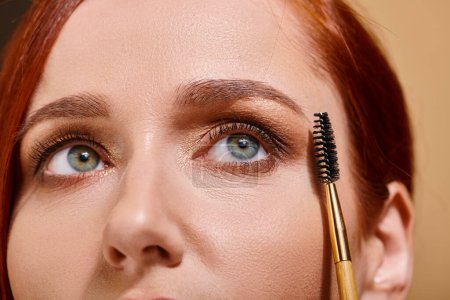 Photo for Cropped photo of redhead woman with green eyes holding eye brow brush on beige backdrop - Royalty Free Image