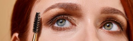 close up photo of redhead woman with green eyes applying mascara on beige background, banner