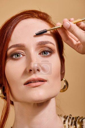portrait of redhead woman with green eyes holding eye brow brush on beige background, makeup