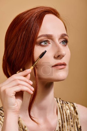 portrait of redhead woman with green eyes applying mascara on beige background, makeup beauty