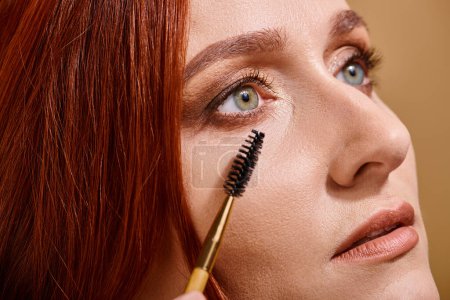 close up view of redhead woman with green eyes applying mascara on beige background, makeup