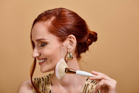 happy redhead woman in gold sequin dress holding makeup brush on beige background, beauty gaze