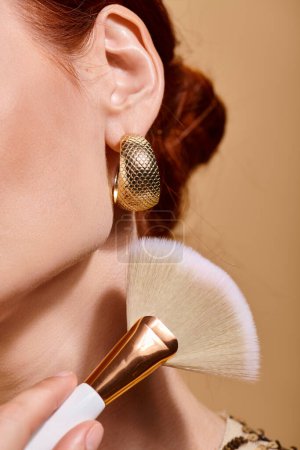 mujer pelirroja recortada en pendiente de oro aplicando bronceador con cepillo de maquillaje sobre fondo beige
