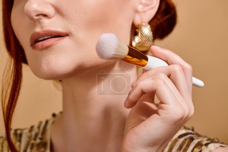 geschnittene rothaarige Frau in goldenem Ohrring mit Make-up-Pinsel für flüssige Foundation-Applikation
