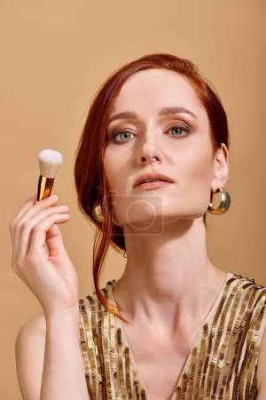 selbstbewusste rothaarige Frau in goldenen Ohrringen mit Make-up-Pinsel auf beigem Hintergrund, Schönheitskonzept
