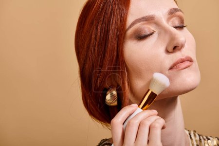 redhead woman in gold earring applying face foundation with makeup brush on beige backdrop