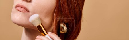 redhead woman in gold earring applying face foundation with makeup brush on beige backdrop, banner
