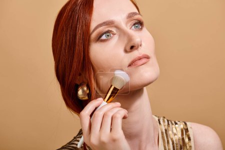 redhead woman in gold earring applying face foundation with makeup brush on beige backdrop