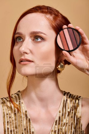 redhead woman in her 30s holding eye shadow palette on beige background, makeup concept