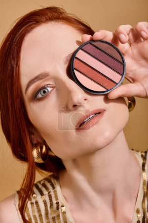 redhead woman covering her face with eye shadow palette on beige background, beauty concept