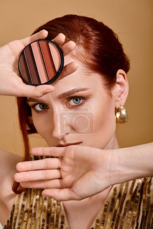 close up of redhead woman holding eye shadow palette above head on beige background, makeup concept