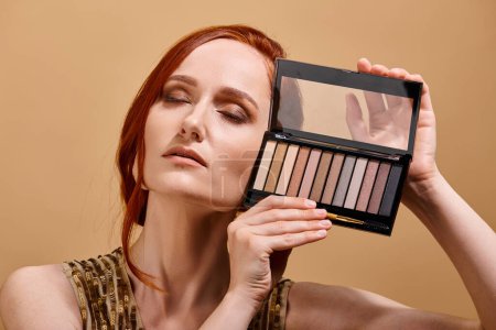 redhead woman holding eye shadow palette near face on beige background, makeup advertisement